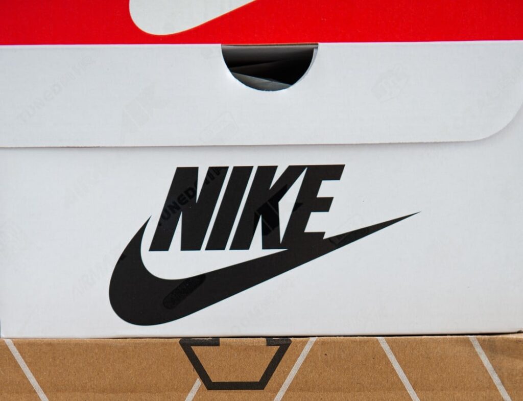 White cardboard shoe box with a black Nike check. It sits on top of a brown shoebox and is below a red shoebox.