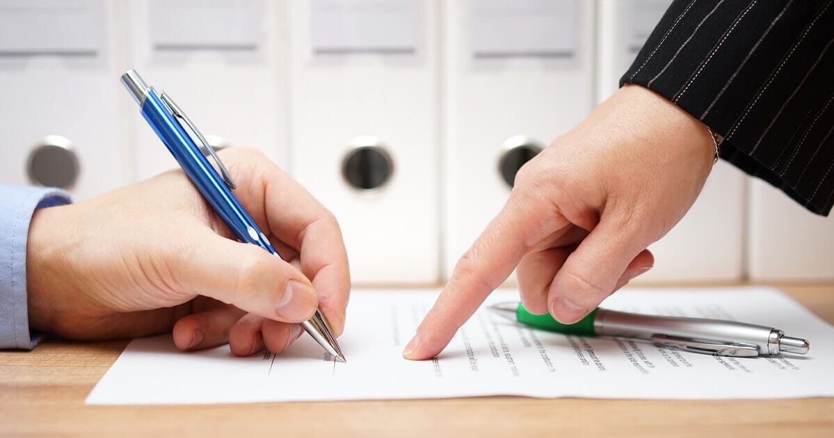 A hand signing a contract with a blue pen. Another hand is pointing to the contract.