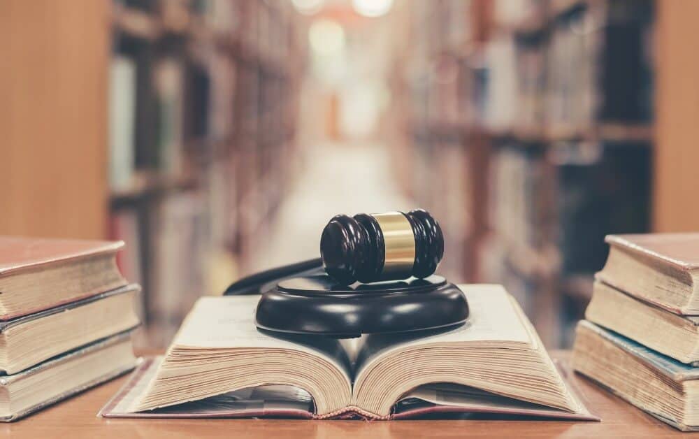 A mahogany wood gavel on top of an open "brand and trademark protection" book with towering bookshelves in a library background