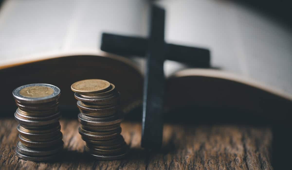 A shadowy cross and two stacks of coins sit in front of an open bible on a walnut desk.